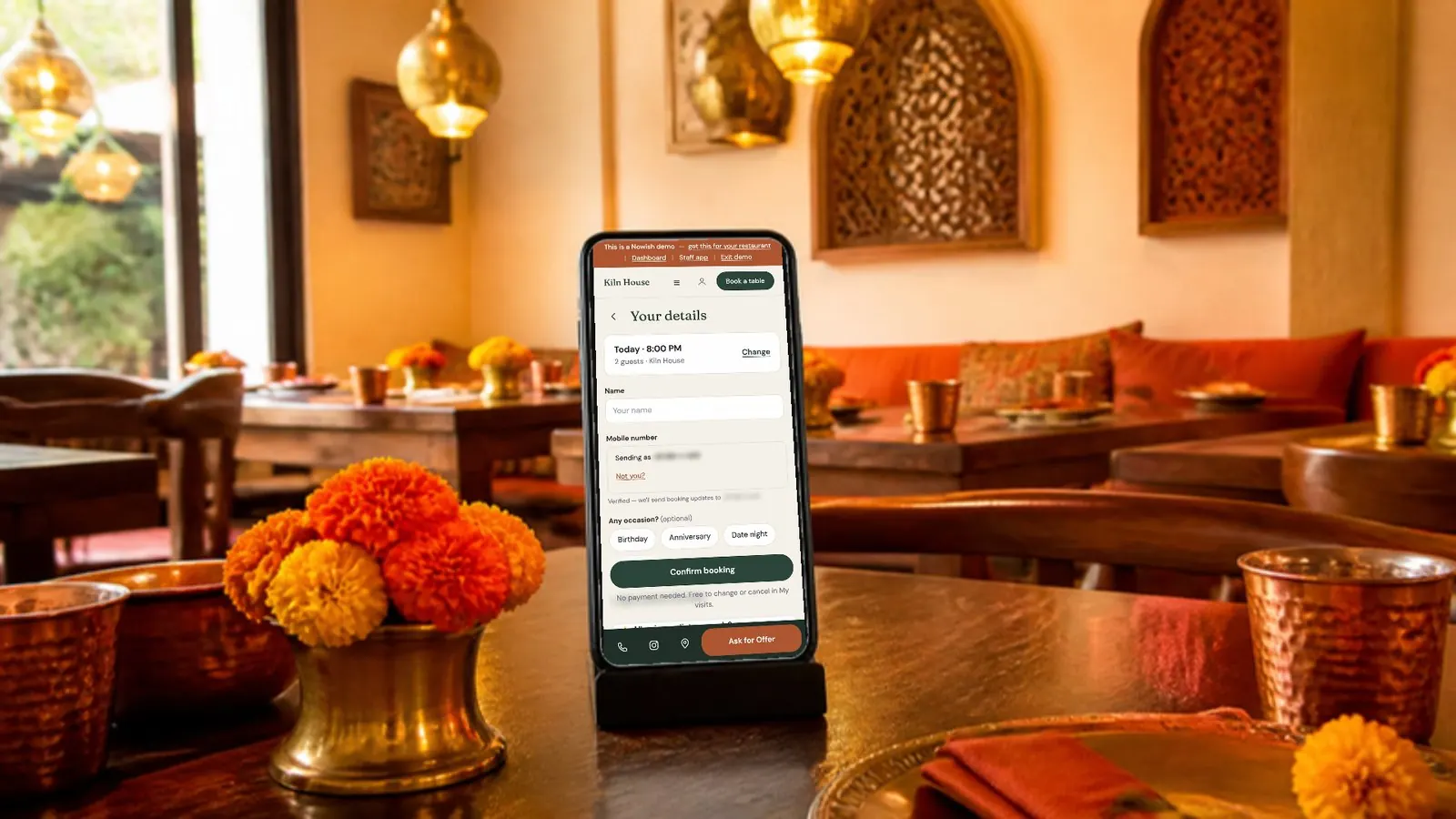 A warm restaurant dining room with marigolds on the table and a phone showing a table being booked on the restaurant's own website.